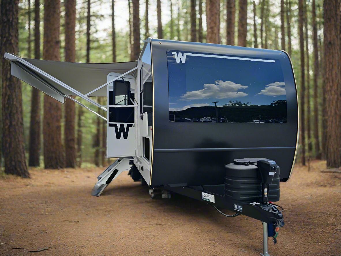 Black travel trailer with a white W logo parked on a paved area with grass and trees in the background.